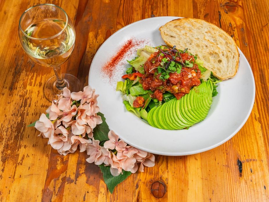 Rustic wooden table setting with a vibrant mixed-greens salad topped with diced tomato salsa and microgreens, fanned avocado slices and a slice of sourdough, paired with a glass of white wine and pale pink hydrangea.