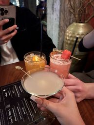 Three craft cocktails raised in a toast at a cozy bar — frosted martini in front and two garnished highballs (orange slice, watermelon skewer) on a wooden table next to a menu and phone, nightlife scene.