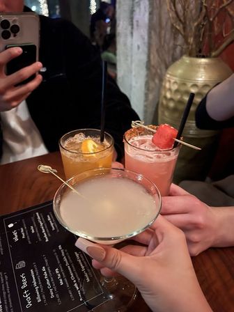 Three craft cocktails raised in a toast at a cozy bar — frosted martini in front and two garnished highballs (orange slice, watermelon skewer) on a wooden table next to a menu and phone, nightlife scene.