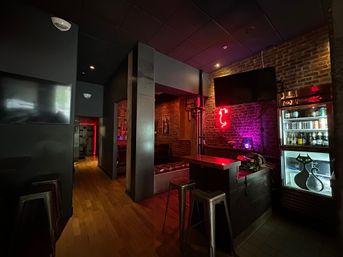 Cozy urban bar interior with exposed brick, wooden floors, metal stools, a small counter, glowing red neon cat sign and a glass-front fridge stocked with drinks.