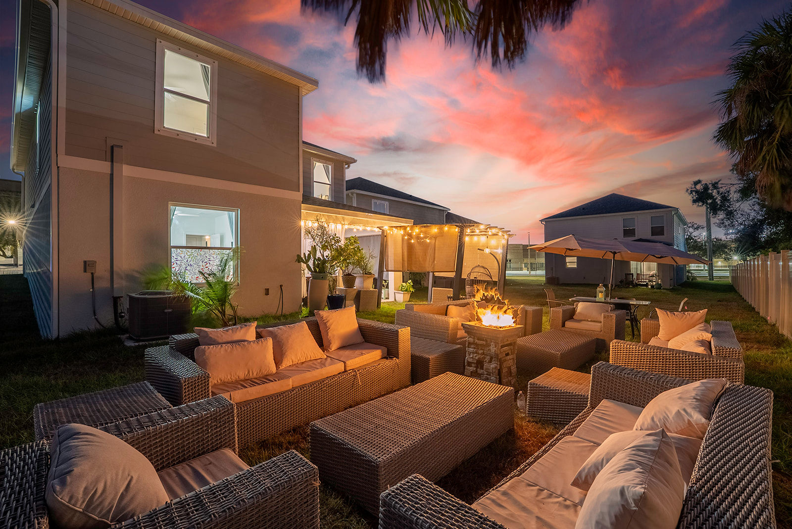 Sunset-lit suburban backyard patio with wicker sofas and cushions arranged around a glowing fire pit, string lights draped over a pergola, umbrellas and potted plants beside a two-story house and palm trees