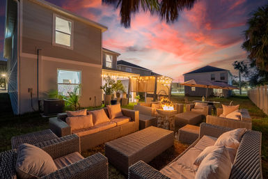 Sunset-lit suburban backyard patio with wicker sofas and cushions arranged around a glowing fire pit, string lights draped over a pergola, umbrellas and potted plants beside a two-story house and palm trees