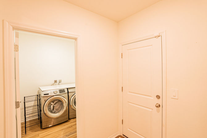 Bright laundry nook off an entryway with stainless front-load washer and dryer, white walls, wood-look flooring and a black metal laundry rack.