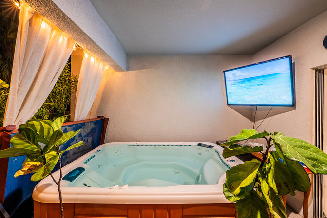 Cozy covered balcony with a private hot tub/jacuzzi, tropical potted plants, sheer curtains and warm string lights, plus a wall-mounted TV showing a sunny beach scene for resort-style relaxation.
