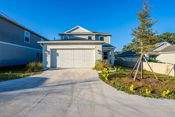Sunlit suburban two-story home with a white two-car garage, concrete driveway, small landscaped front yard with a staked young tree and ornamental grasses under a clear blue sky.