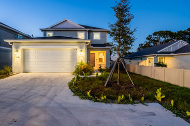 Two-story suburban home at dusk with a glowing two-car garage, warm front-porch light, concrete driveway and landscaped yard with a young staked tree and low plants.