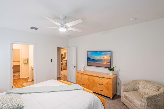 Bright modern primary bedroom with white ceiling fan, bed with white bedding, light wood dresser topped with a potted plant and sun sculpture, wall-mounted TV showing a beach sunset, cozy beige armchair, and open doors to an en-suite bathroom and hallway.