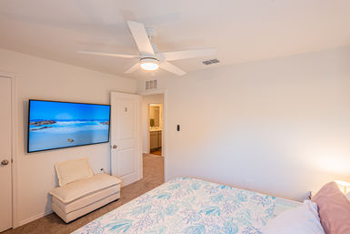 Bright coastal-themed bedroom with white ceiling fan, wall-mounted TV showing a beach, cream ottoman, floral blue-and-white bedding and open door to bathroom.