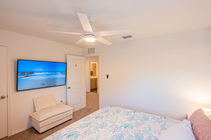 Bright coastal-themed bedroom with white ceiling fan, wall-mounted TV showing a beach, cream ottoman, floral blue-and-white bedding and open door to bathroom.