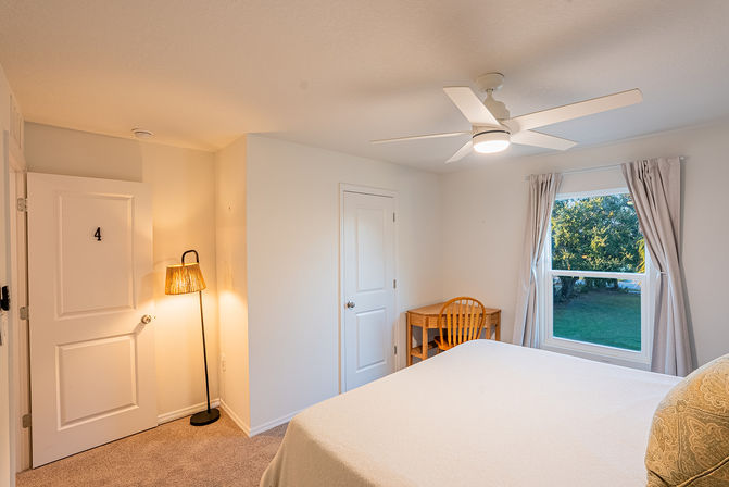 Cozy bright bedroom with white ceiling fan, neutral bedding, a floor lamp and small wooden desk and chair by a window overlooking a green yard and trees.