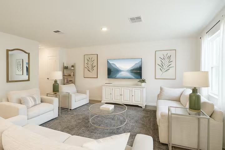 Bright airy living room with white sofas and armchairs around a round glass coffee table on a gray rug, wall-mounted TV showing a mountain scene, white media console, botanical prints, green table lamps and a large window letting in natural light.
