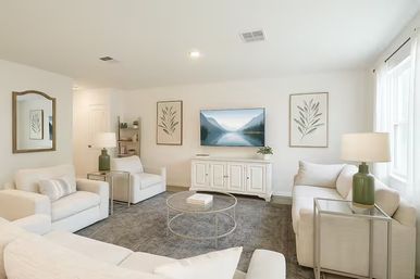 Bright airy living room with white sofas and armchairs around a round glass coffee table on a gray rug, wall-mounted TV showing a mountain scene, white media console, botanical prints, green table lamps and a large window letting in natural light.