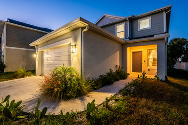 Twilight exterior of a modern two-story suburban house with illuminated garage, warm-lit front entry, concrete driveway and tropical landscaping