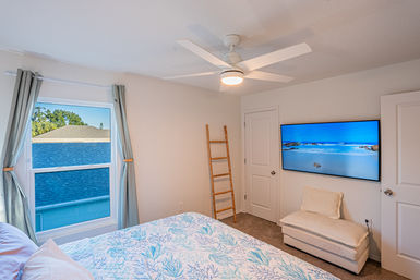 Cozy coastal-themed bedroom with white ceiling fan, large curtained window, wall-mounted TV showing a beach scene, rustic wooden ladder décor, upholstered bench, and blue coral-pattern quilt on the bed.