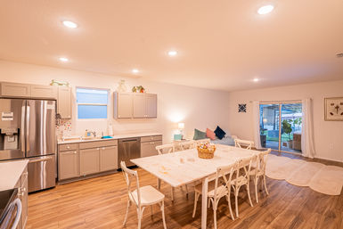 Bright open-plan kitchen and dining area with distressed white farmhouse table and chairs, gray shaker cabinets, stainless-steel refrigerator, hardwood floors, cozy sofa with colorful pillows, and sliding glass door to a patio.