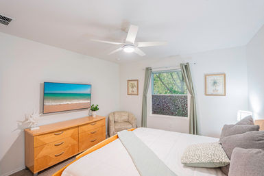 Bright modern bedroom with white ceiling fan, light wood dresser, wall-mounted TV showing a beach scene, window with green curtains and cozy neutral bedding for an airy coastal vibe.
