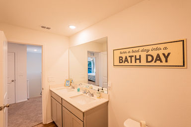 Bright neutral residential bathroom with double-sink vanity, large mirror, upbeat wall sign reading 'turn a bad day into a BATH DAY', toilet and doorway to a carpeted bedroom visible