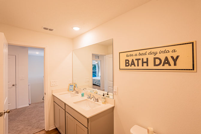 Bright neutral residential bathroom with double-sink vanity, large mirror, upbeat wall sign reading 'turn a bad day into a BATH DAY', toilet and doorway to a carpeted bedroom visible