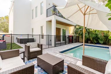 Sunny modern two-story home backyard with a fenced rectangular pool, wicker patio seating and umbrella on a paver deck, tropical plants and hot tub.