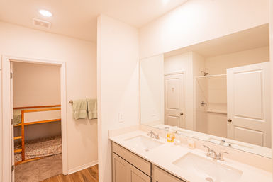 Bright modern family bathroom with white double-sink vanity and large mirror, walk-in tiled shower, green hand towels and wood-look floor, doorway opening to a bunk-bed bedroom — ideal for family or rental stays.