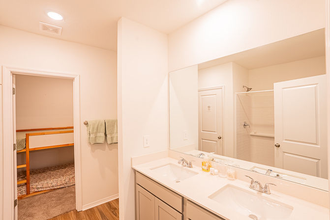 Bright modern family bathroom with white double-sink vanity and large mirror, walk-in tiled shower, green hand towels and wood-look floor, doorway opening to a bunk-bed bedroom — ideal for family or rental stays.