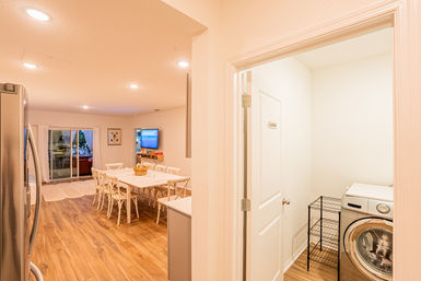 Bright open-plan kitchen and dining area with white farmhouse table and chairs, wall-mounted TV and sliding glass door to patio; adjacent laundry closet with front-load washer and wire shelving on warm wood floors.