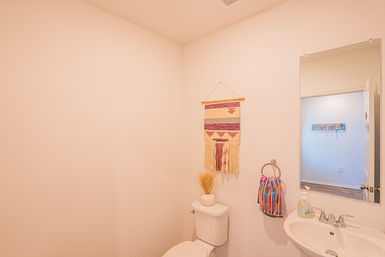 Cozy neutral powder room in a home with a white toilet and pedestal sink, colorful striped hand towel, boho woven wall hanging and a mirror reflecting a hallway.