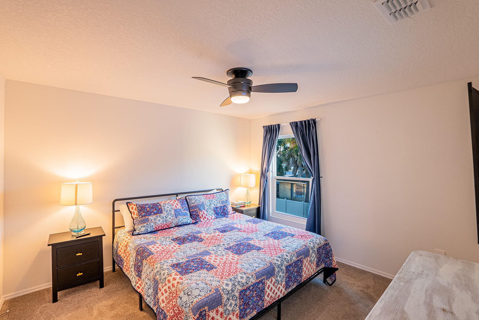 Cozy residential bedroom with colorful patchwork quilt on bed, modern ceiling fan, two nightstands with lamps, navy curtains framing a window with palm trees outside — tidy interior ideal for real estate or vacation rental listing.