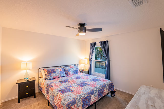 Cozy residential bedroom with colorful patchwork quilt on bed, modern ceiling fan, two nightstands with lamps, navy curtains framing a window with palm trees outside — tidy interior ideal for real estate or vacation rental listing.