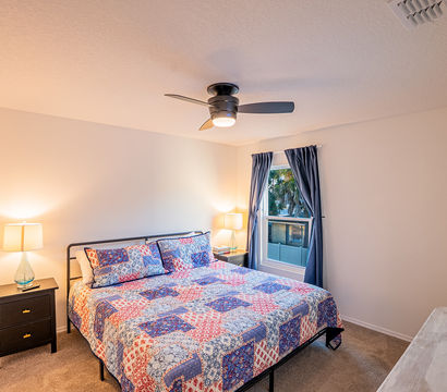 Cozy residential bedroom with colorful patchwork quilt on bed, modern ceiling fan, two nightstands with lamps, navy curtains framing a window with palm trees outside — tidy interior ideal for real estate or vacation rental listing.