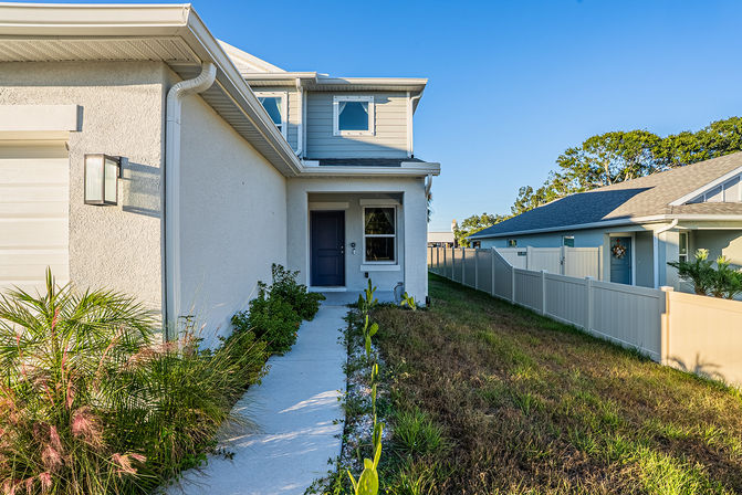 Sunlit front walkway to a modern two-story suburban house with stucco siding and a blue entry door, small palm and garden plants, vinyl privacy fence and neighboring home under a clear blue sky