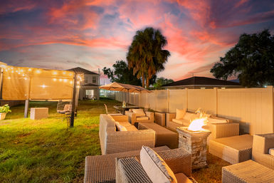 Cozy backyard patio lounge with wicker sofas, glowing fire pit, string lights and umbrella beneath a pink sunset and tall palm tree.