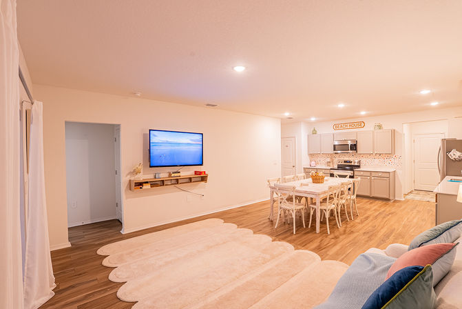 Bright open-plan living room and kitchen with hardwood floors, scalloped beige rug, wall-mounted TV, farmhouse dining table, light-gray cabinets, stainless appliances and beach-house decor.