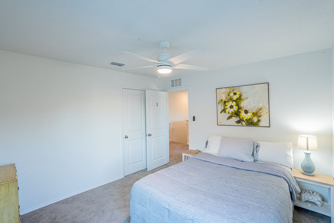 Bright, airy bedroom with gray quilted bed, white ceiling fan, floral wall art, wooden nightstand with blue lamp, carpeted floor and an open closet door.