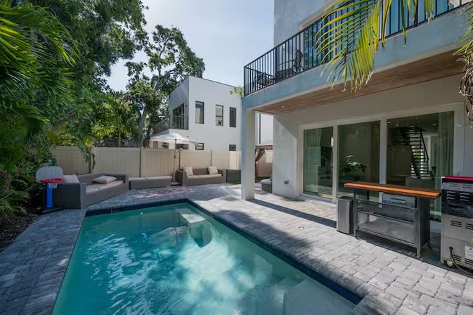 Sunlit modern two-story home with a rectangular backyard pool, stone paver patio, outdoor kitchen island, balcony, palm trees and cushioned lounge seating with umbrella.