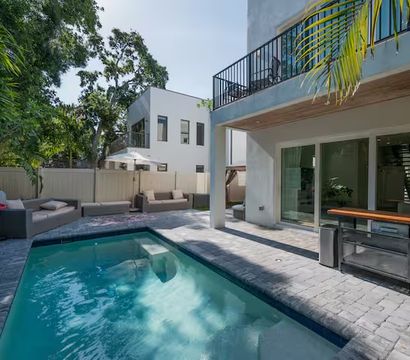 Sunlit modern two-story home with a rectangular backyard pool, stone paver patio, outdoor kitchen island, balcony, palm trees and cushioned lounge seating with umbrella.