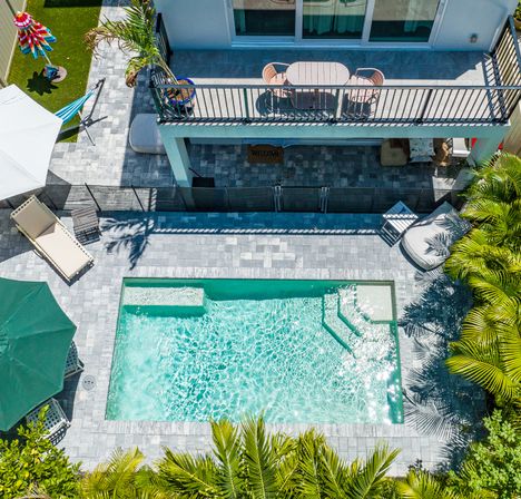 Aerial view of a tropical backyard with a sparkling turquoise rectangular pool, light-gray tiled patio, upper balcony dining set, lounge chairs, colorful umbrella and palm trees casting shadows.