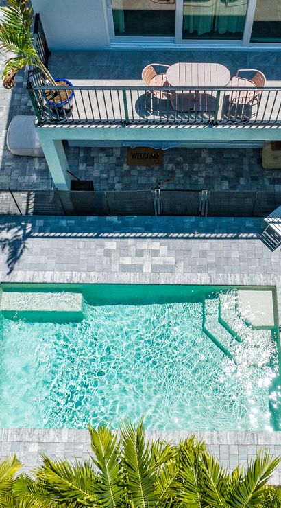 Aerial view of a tropical backyard with a sparkling turquoise rectangular pool, light-gray tiled patio, upper balcony dining set, lounge chairs, colorful umbrella and palm trees casting shadows.
