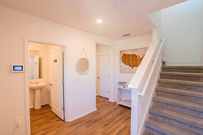 Bright residential entryway with wood-look floors, carpeted staircase, round hanging mirror, small console table, wall-mounted smart thermostat, open half-bath with pedestal sink, and rustic wooden map of the United States.