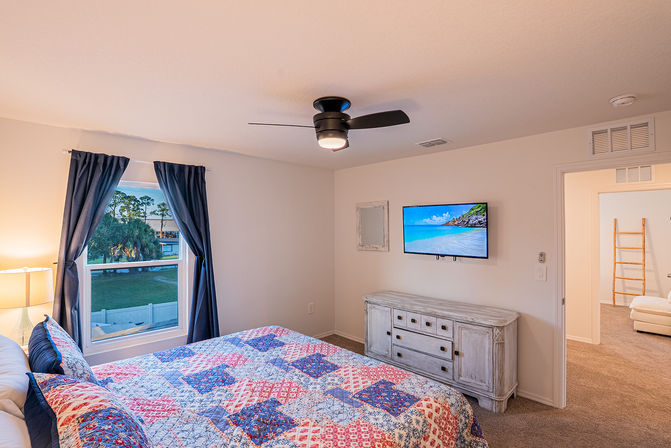 Cozy bedroom with modern ceiling fan, navy curtains framing a window with palm trees outside, colorful patchwork quilt on the bed, whitewashed dresser and wall-mounted TV showing a beach scene.