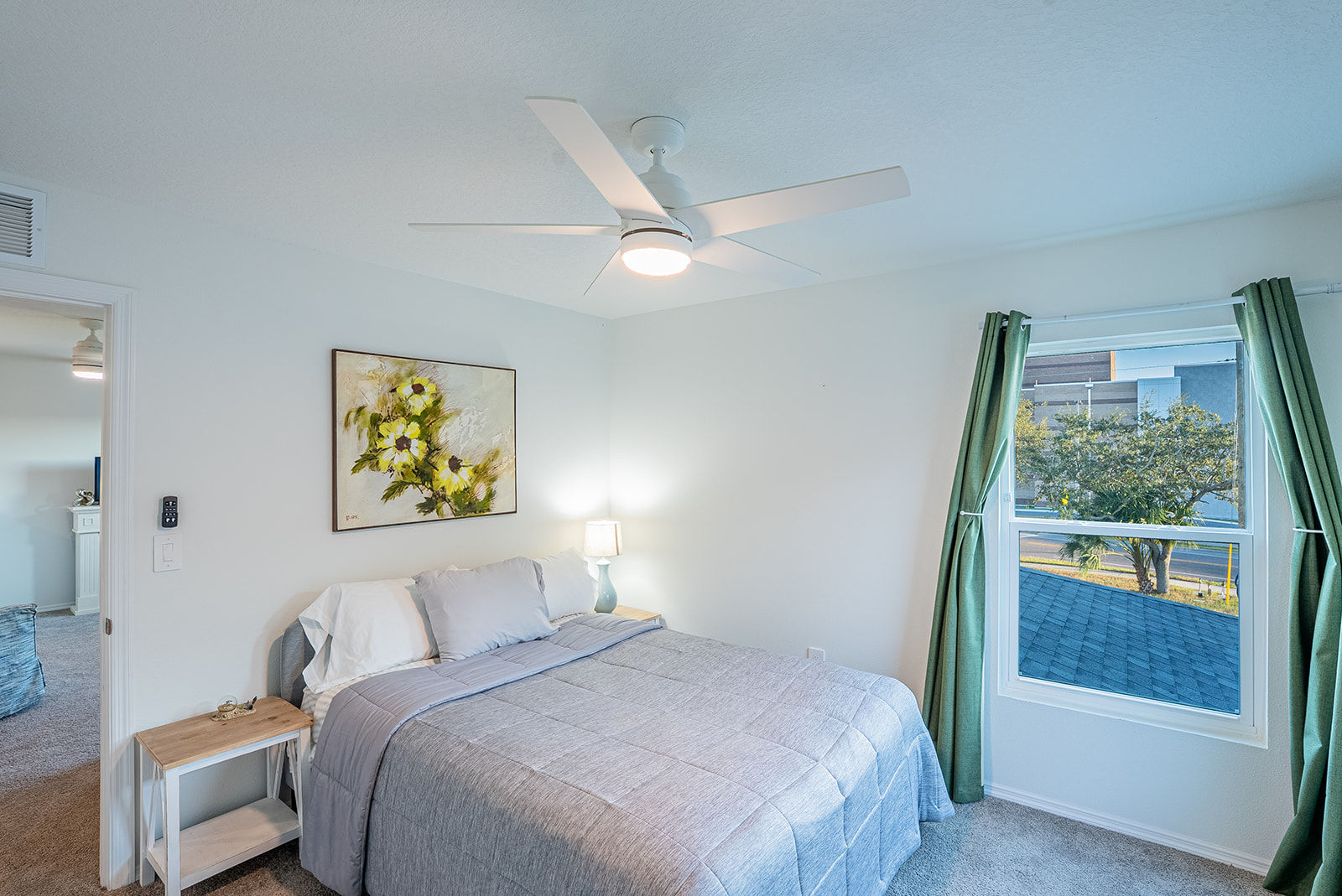 Bright, cozy bedroom with gray quilted bed, white ceiling fan and light, floral wall art, bedside lamp on a small wooden table, and a window with green curtains overlooking a tree-lined suburban street.