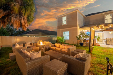 Cozy suburban backyard patio at sunset with wicker outdoor seating arranged around a glowing stone fire pit, string lights on a pergola, potted plants, and a two-story house in the background.