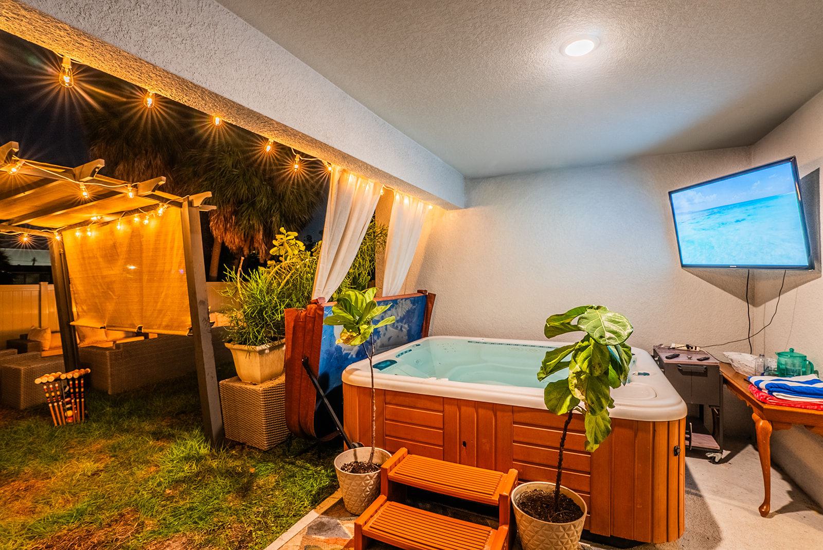 Cozy backyard patio at night with a wooden hot tub and orange steps, potted plants, warm string lights draping a pergola and curtained seating area, plus a wall-mounted TV showing an ocean scene.