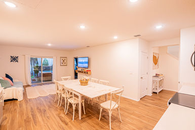 Bright, airy open-plan dining and living room in a modern suburban home — light wood floors, white farmhouse dining table with eight chairs, wall-mounted TV showing a beach scene, sliding glass doors to a backyard patio and a cozy sofa with soft rugs.