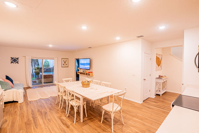 Bright, airy open-plan dining and living room in a modern suburban home — light wood floors, white farmhouse dining table with eight chairs, wall-mounted TV showing a beach scene, sliding glass doors to a backyard patio and a cozy sofa with soft rugs.