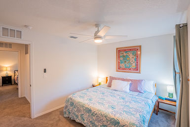 Cozy coastal-themed bedroom with king-size bed, blue-and-white seaweed quilt, pillows, ceiling fan, framed map art above bed, bedside lamps, carpeted floor and curtained window