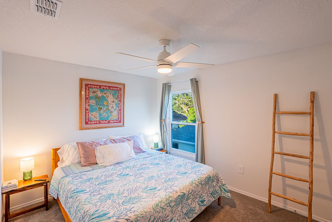 Bright coastal-style bedroom with queen bed, sea-themed quilt, bedside lamps, framed Florida map, white ceiling fan, and decorative wooden ladder