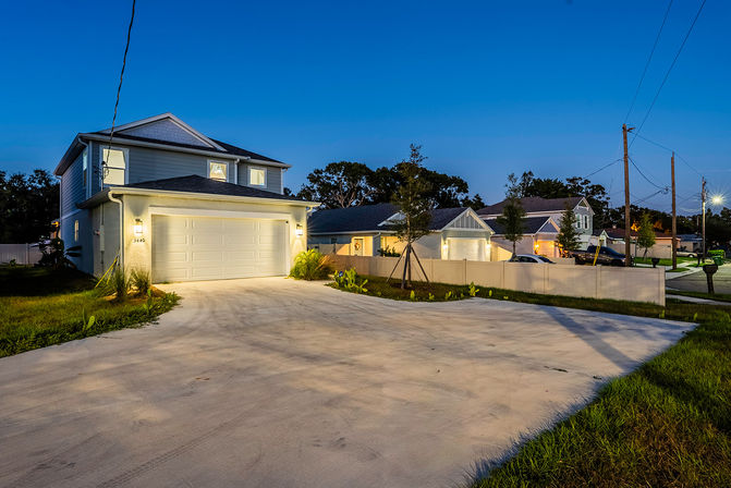 Twilight view of a modern two-story suburban home with a lit double garage, wide concrete driveway, young trees and fences lining a quiet neighborhood street under a deep blue sky.