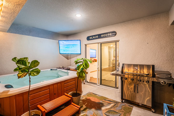 Cozy covered beach-house patio with a hot tub, wall TV, potted tropical plants, and stainless-steel grill beside sliding glass doors.