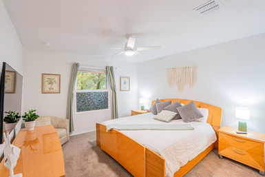 Bright, cozy bedroom with light wood platform bed, white bedding and gray throw pillows, macramé wall hanging, ceiling fan, matching wood nightstands with green lamps and a window overlooking leafy trees